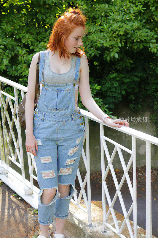 这是一个14 / 15岁的快乐微笑的少女，留着红色短发/姜黄色短发，穿着蓝色牛仔裤和灰色运动胸罩，站在河边公园花园的桥上，提着手提包，露出破洞的膝盖